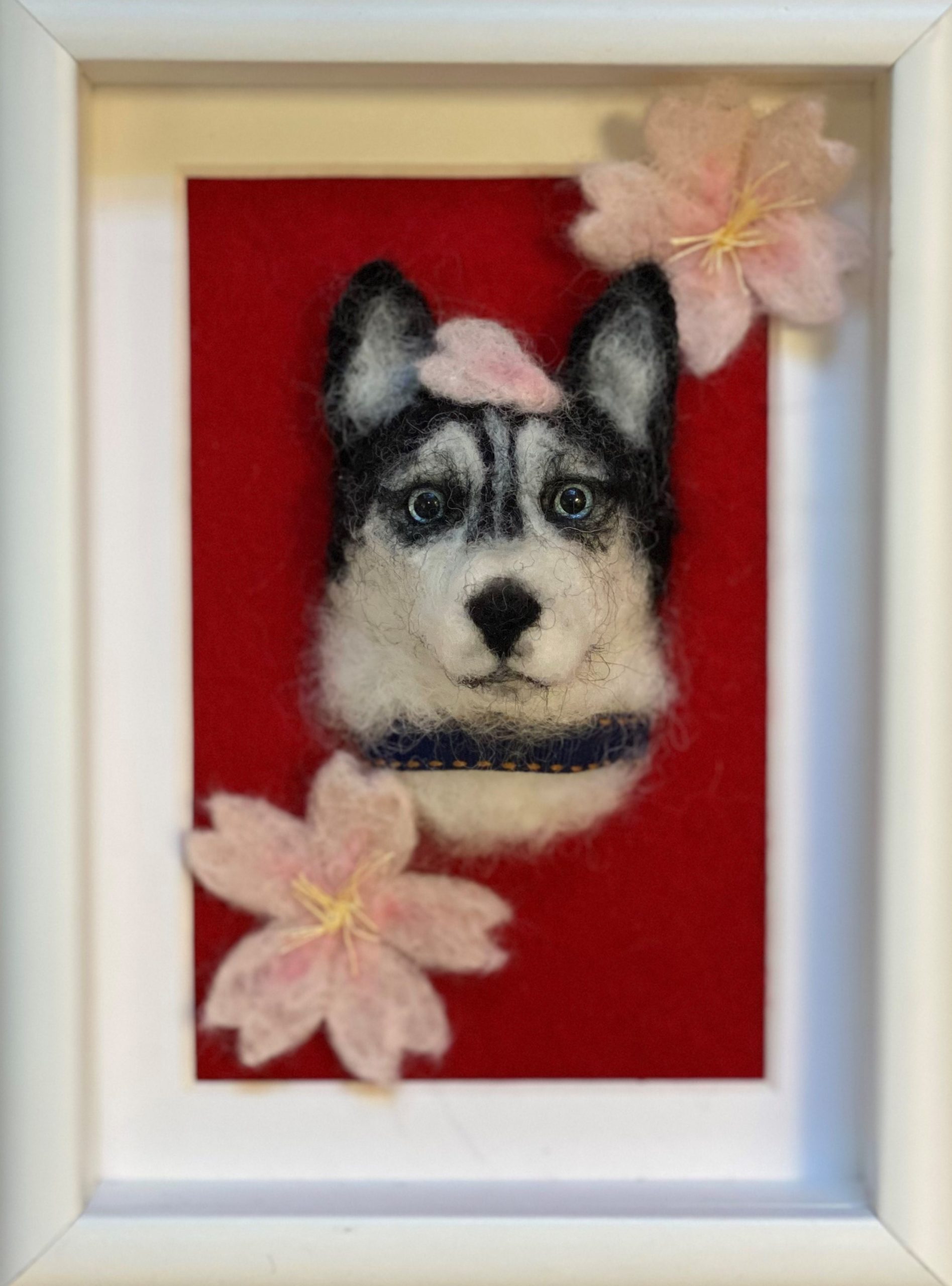 Framed, needle-felted portrait of a husky with 2 pink-and-white flowers in the corners and a flower petal on the huskey's head. The background of the portrait is red and the frame is white. The huskey has blue eyes.