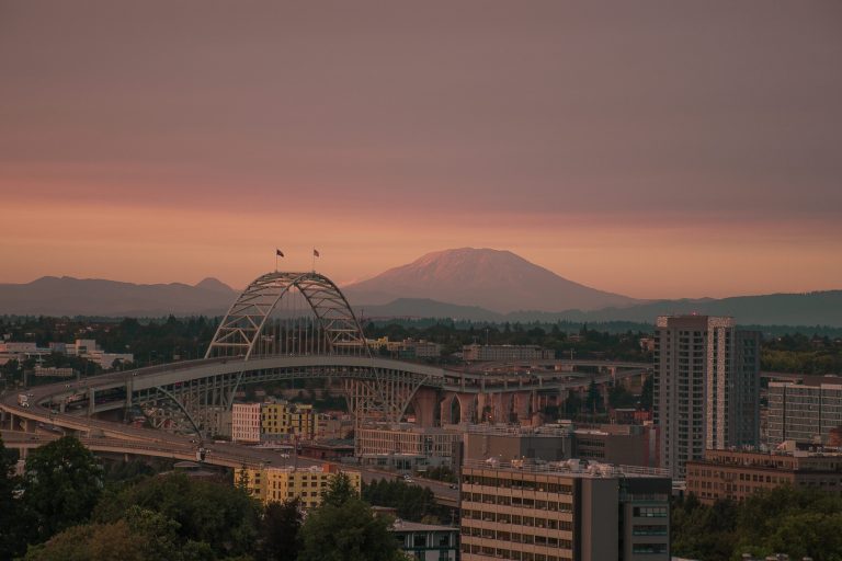 A photograph of a hazy Portland sunset. In the far background is Mount St. Helens. Fremont bridge can be seen, as well as a view of downtown from the South